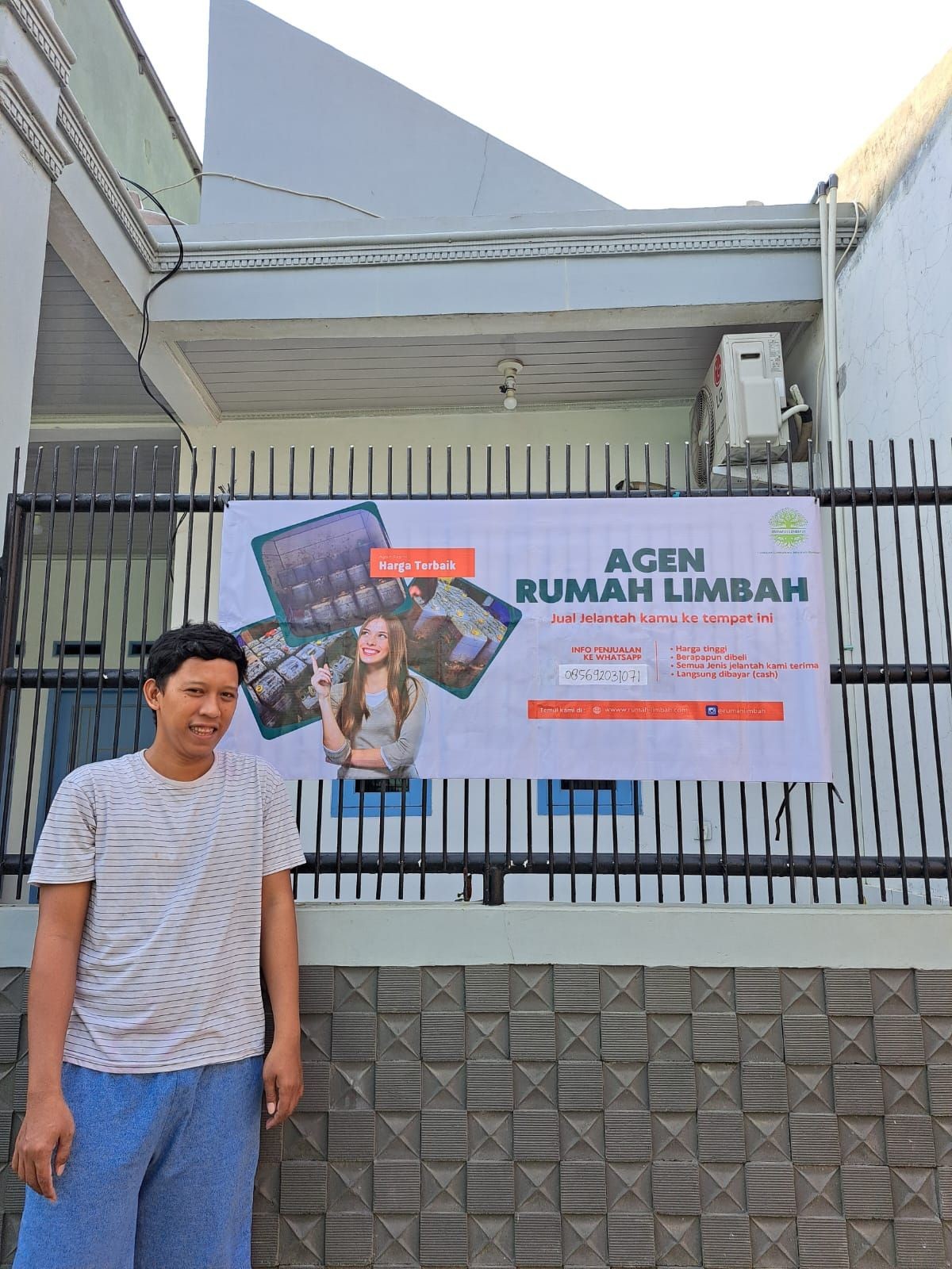 Man standing next to a banner promoting waste recycling in front of a building with air conditioning unit.