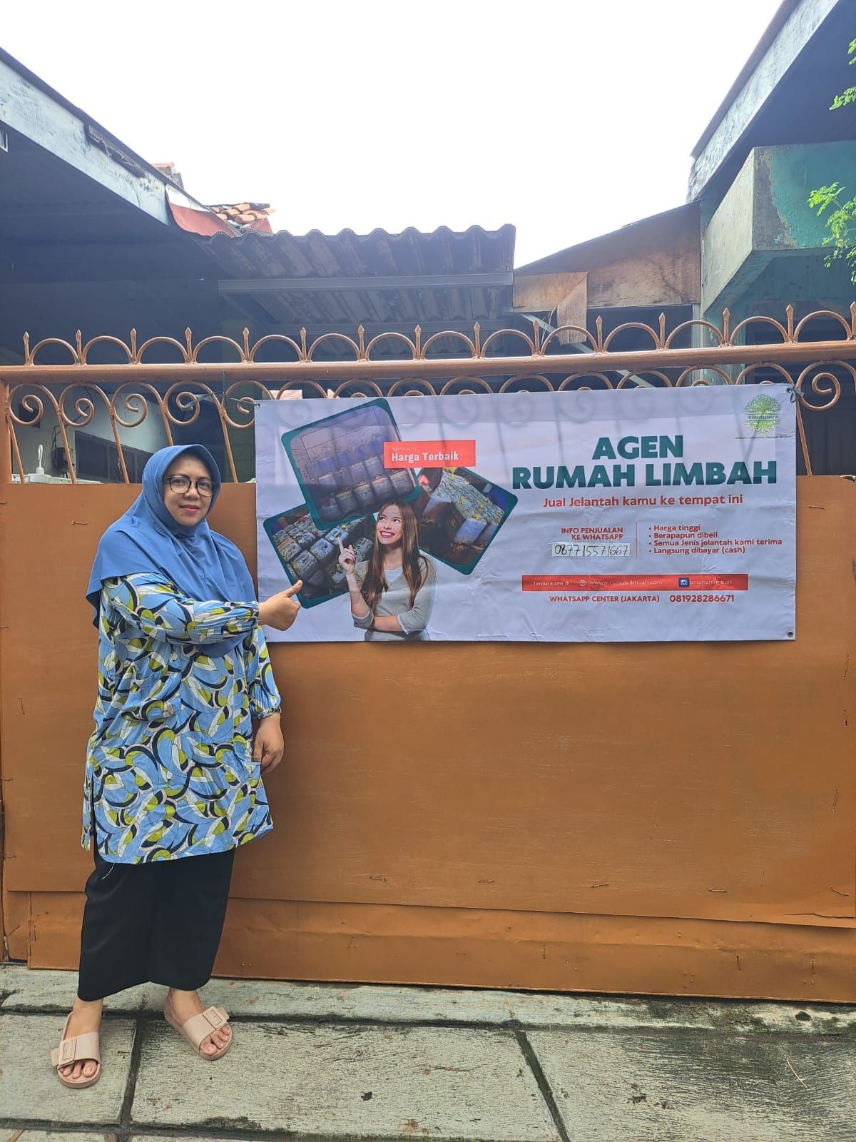 Person in blue hijab pointing to a banner about an oil waste agent on a brown fence.